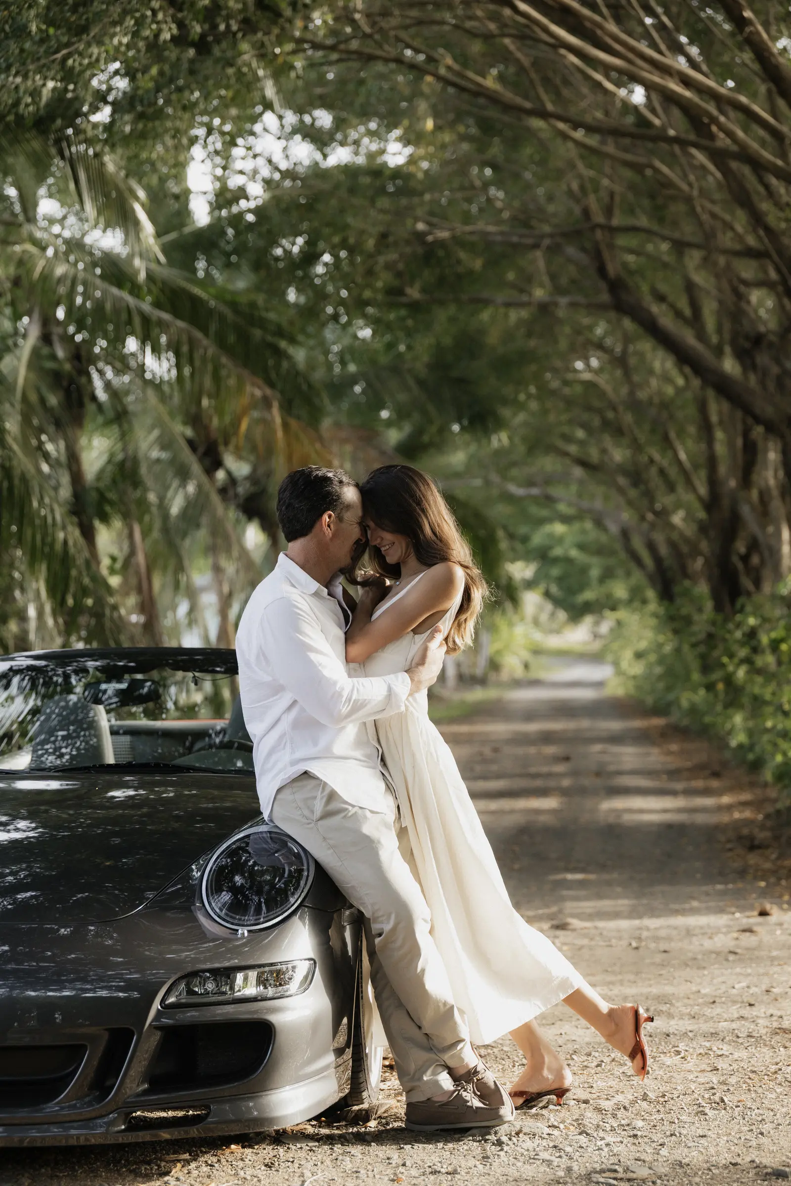 Bride and groom embracing beside a car under trees
