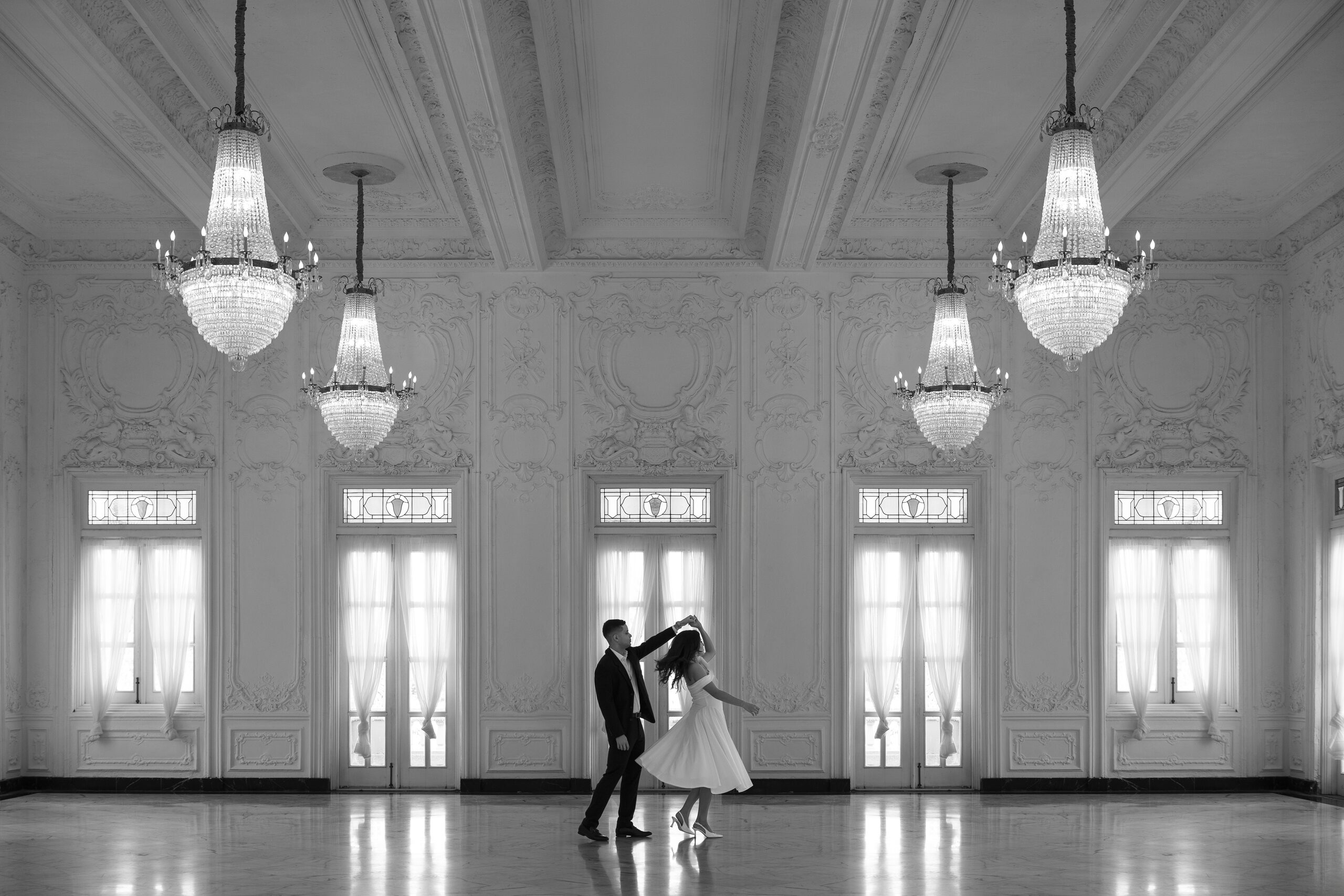 Bride and groom dancing in an ornate ballroom with crystal chandeliers and tall arched windows