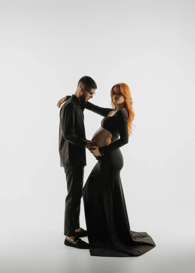 Couple in a black studio portrait, hands resting on the pregnant belly