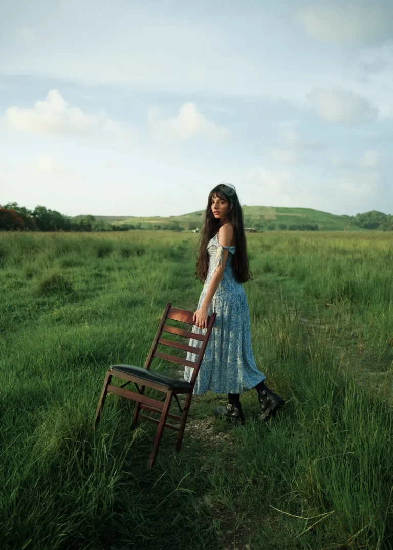 Woman in a blue dress beside a wooden chair in an open grassy field