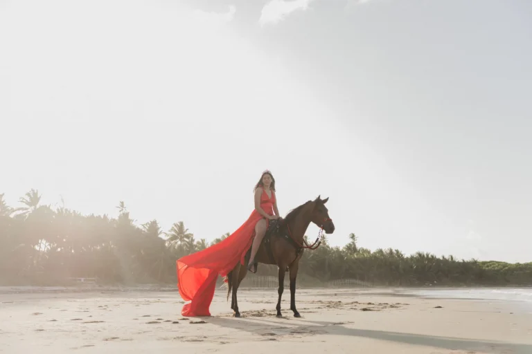 Woman in a flowing red dress riding a horse on the beach at sunset