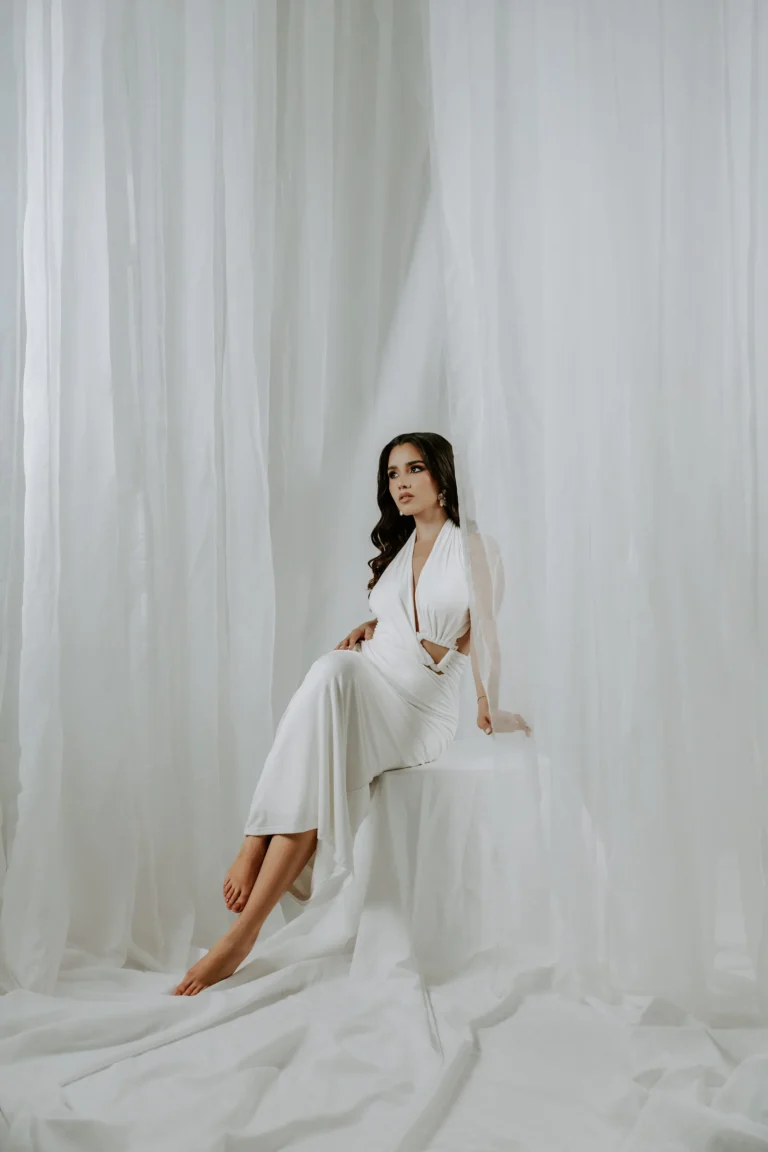 Woman in a white outfit seated in front of sheer curtains