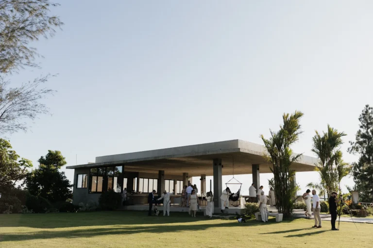 Modern open-air event pavilion with a concrete roof and palm trees, guests gathered underneath